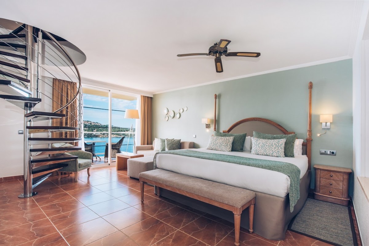 Spacious resort bedroom with a queen bed, spiral staircase, and a balcony overlooking the ocean and marina.