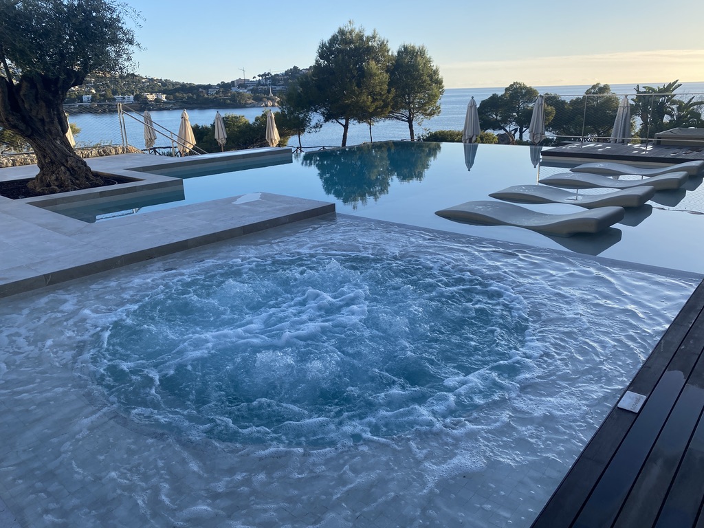 Outdoor jacuzzi with bubbling water in foreground, infinity pool beyond, lounge chairs and umbrellas lining the deck, Mediterranean Sea and coastline visible in background.