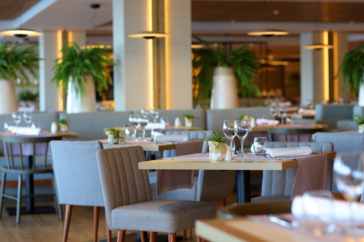Spacious dining room with wooden tables, gray upholstered chairs, place settings, and tall white columns with gold accents beneath warm lighting.