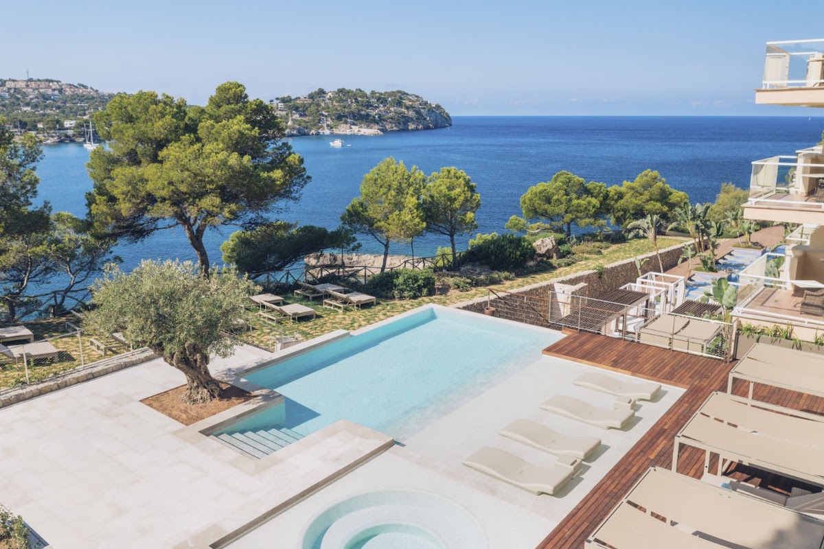 Resort pool with wooden deck and loungers overlooking a blue Mediterranean cove with pine trees and a distant rocky island.