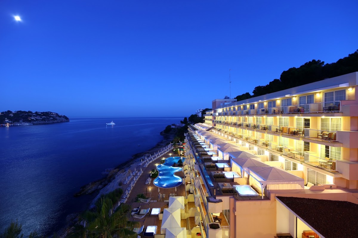 Illuminated cliff-side resort with lit rooms, terraced pool, and lounge chairs overlooking a calm blue sea at dusk.