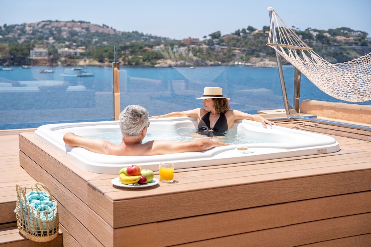 Two guests relax in a beachfront hot tub on a wooden deck overlooking a blue Mediterranean bay with sailboats and a hammock nearby.