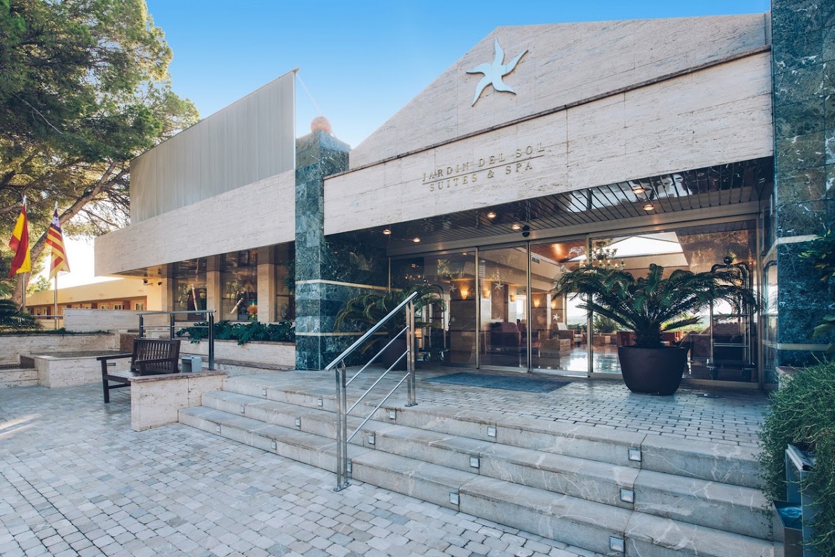Modern entrance to Iberostar Selection Jardín del Sol Suites with stone facade, sun logo, stepped plaza, and landscaped planters under clear sky.