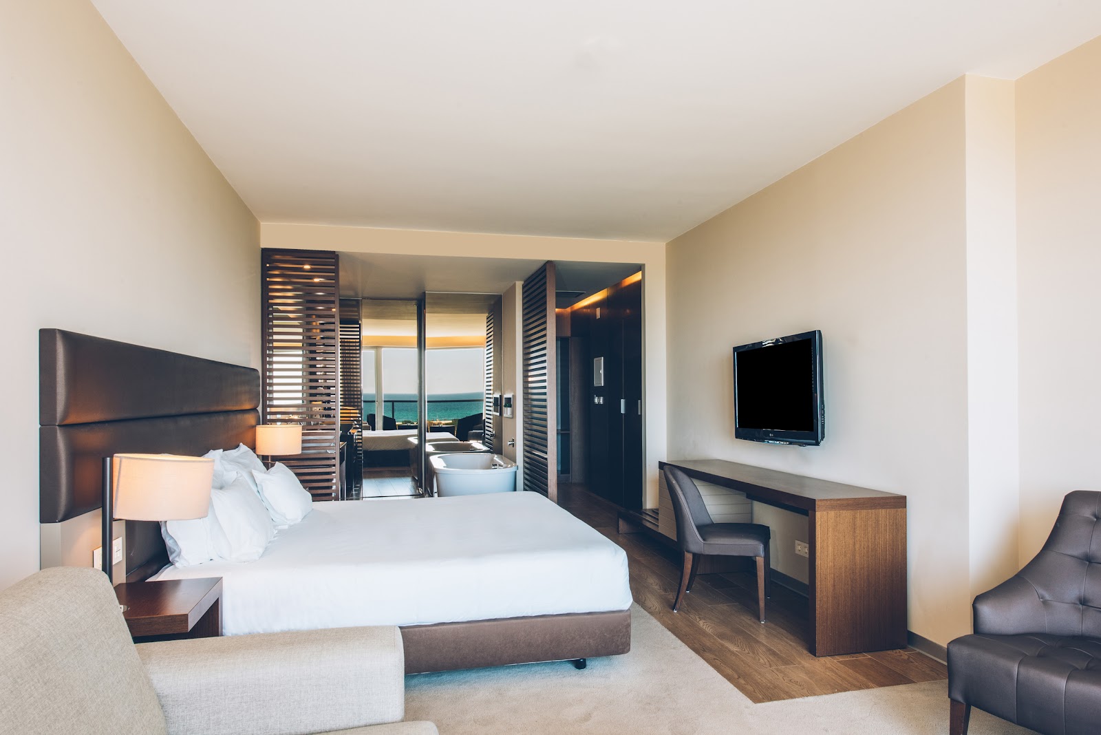 Modern resort suite with soaking tub, dual vanities, and bed with brown upholstered headboard separated by wooden louvered partition.