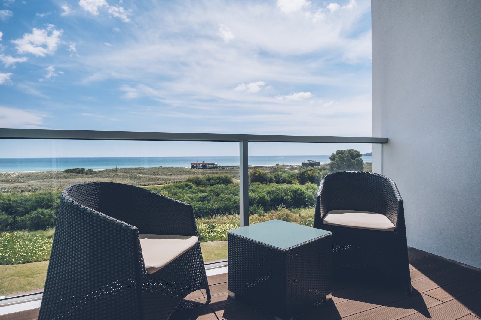 Two black woven chairs with cushions face a table on a terrace overlooking coastal dunes, vegetation, and ocean under blue sky.