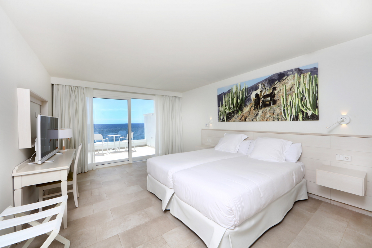 Resort room with queen bed, wood headboard, white linens, olive green armchair, and desert cactus artwork on the wall.