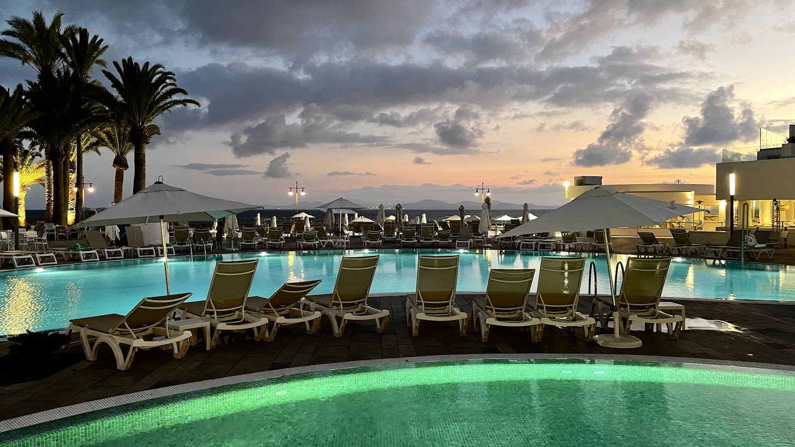 Illuminated pool deck at dusk with loungers, palm trees, and mountain views at Iberostar Selection Lanzarote Park.
