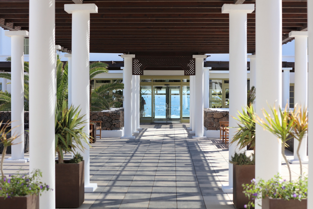 Pool deck with white loungers, blue umbrellas, and dining tables at Iberostar Selection Lanzarote Park, with palm trees and white buildings under clear sky.