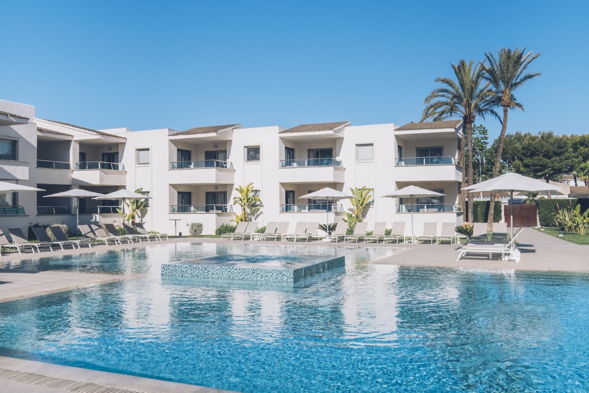 White resort buildings with balconies overlook a large pool with a sunken platform, surrounded by loungers and umbrellas under clear blue sky.