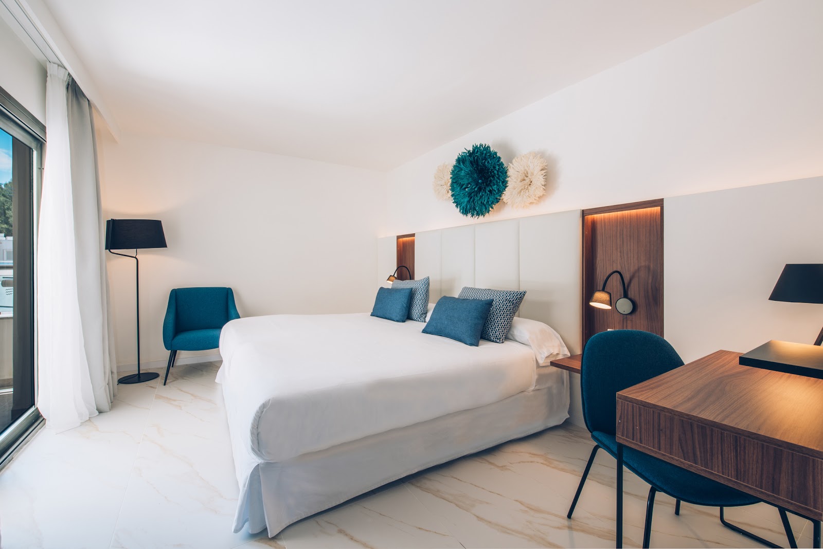 Modern resort bedroom with white bed, teal accent pillows, blue chair, wooden desk, and decorative wall art above the headboard.