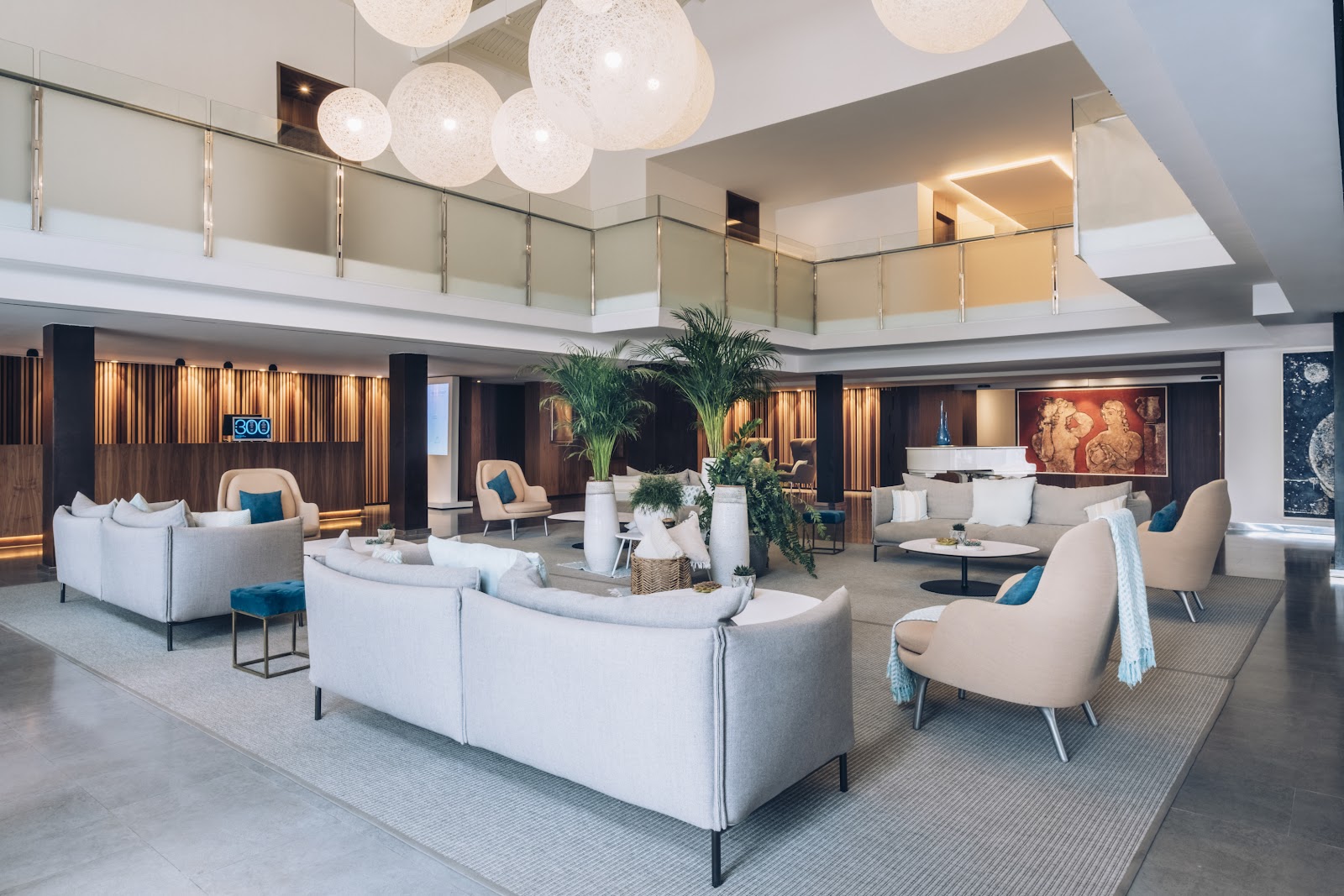 Modern resort lobby with cream sofas, beige armchairs, potted palms, pendant lights, and two-story glass railings overlooking the space.