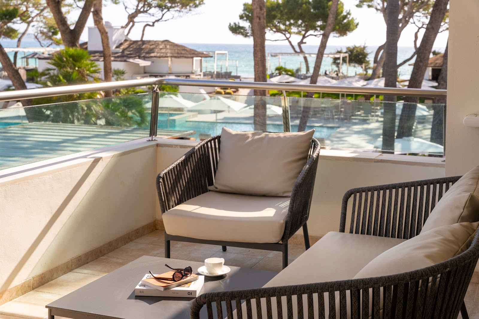 Lounge chairs with cream cushions on a seaside terrace overlooking a pool and Mediterranean beach, with pine trees framing the view.