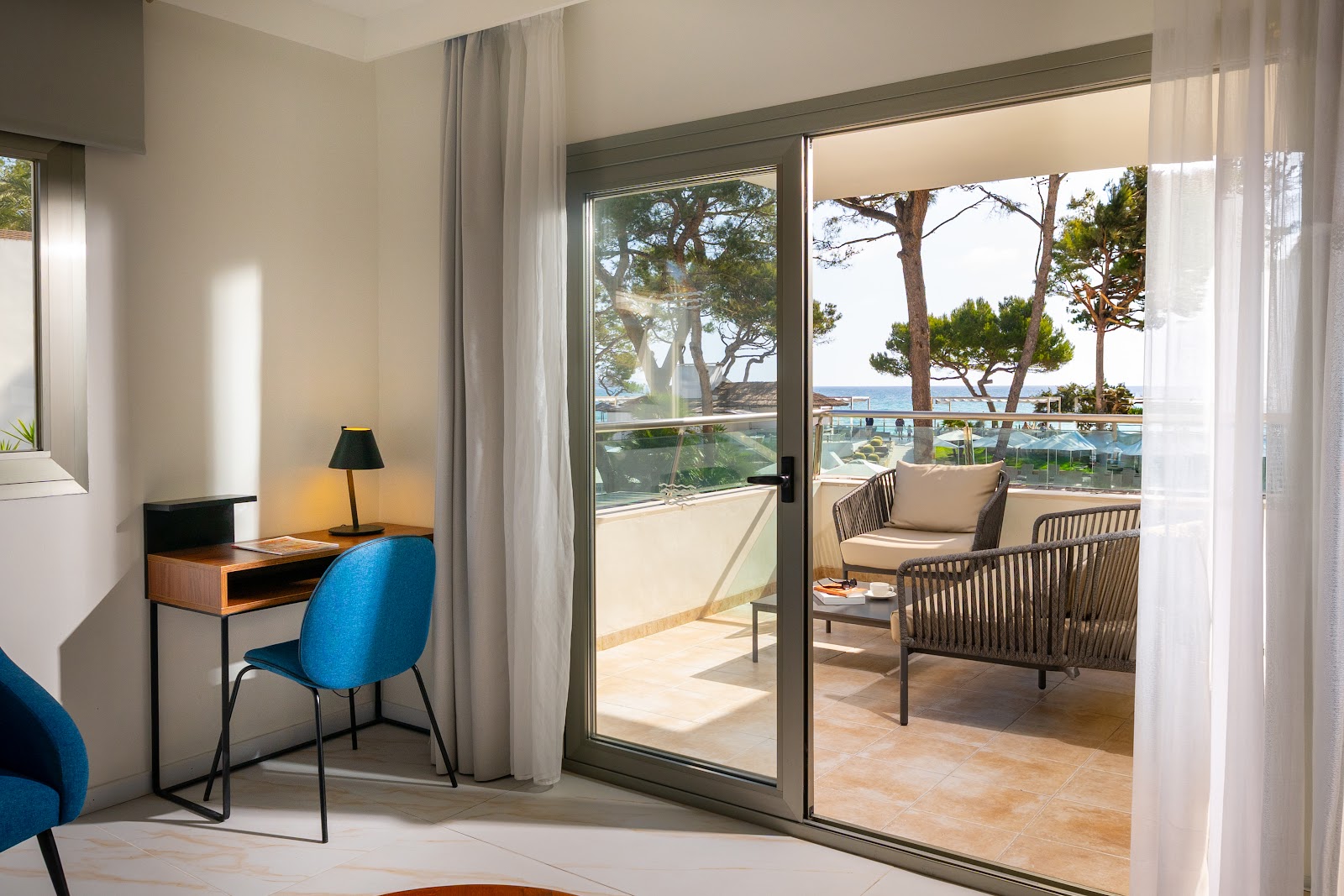 Resort room with desk and blue chair opening onto a terrace with wicker chairs overlooking pine trees and Mediterranean Sea.