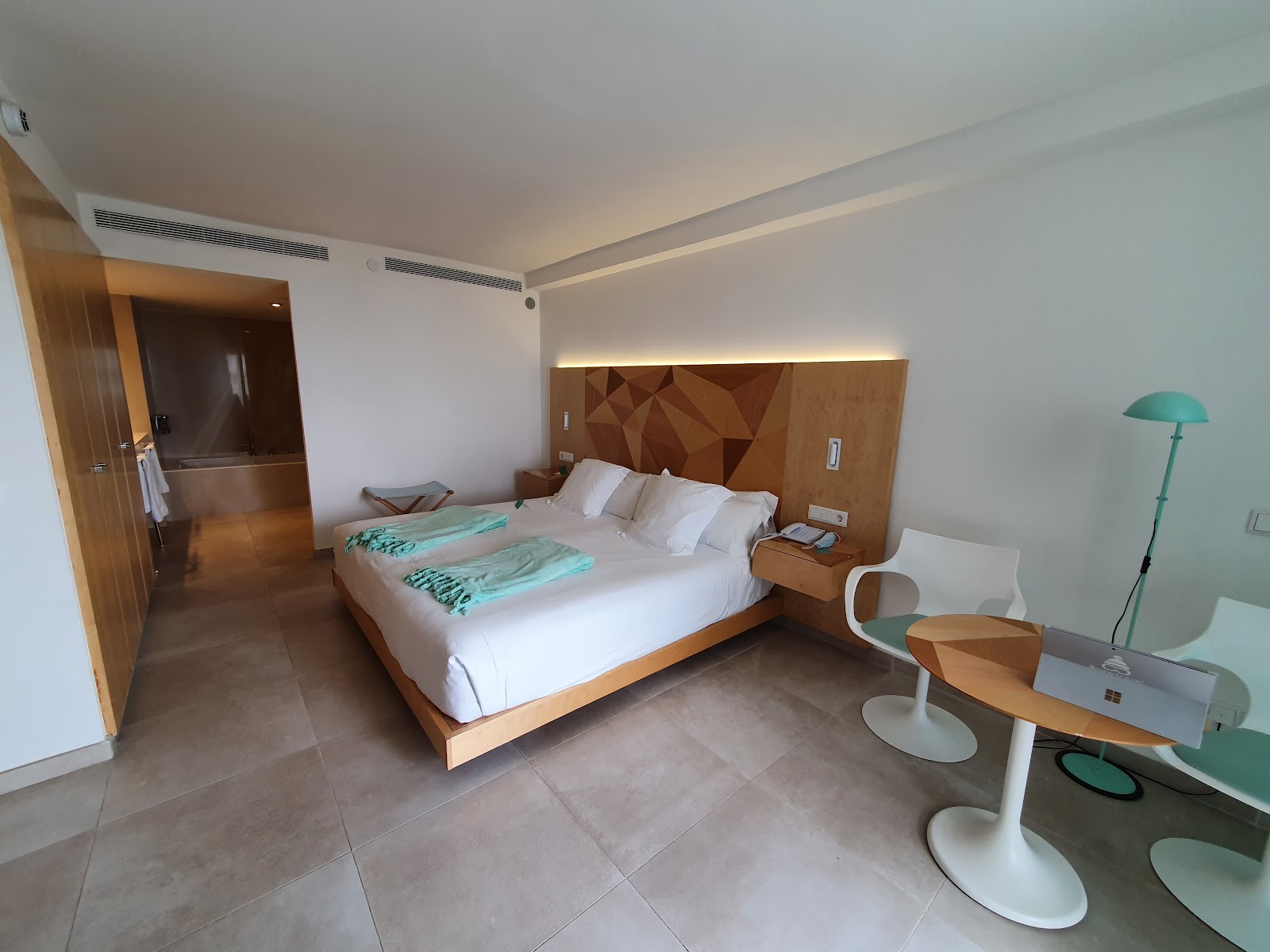 Resort room with wooden accent wall, turquoise curtains, and balcony overlooking a turquoise sea and sandy beach below.