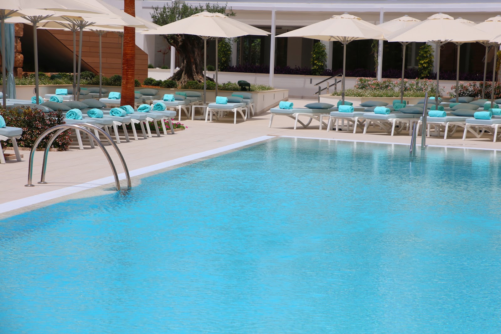 Turquoise pool with metal entry ladders, surrounded by white lounge chairs with turquoise towels, white umbrellas, and modern resort buildings in the background.