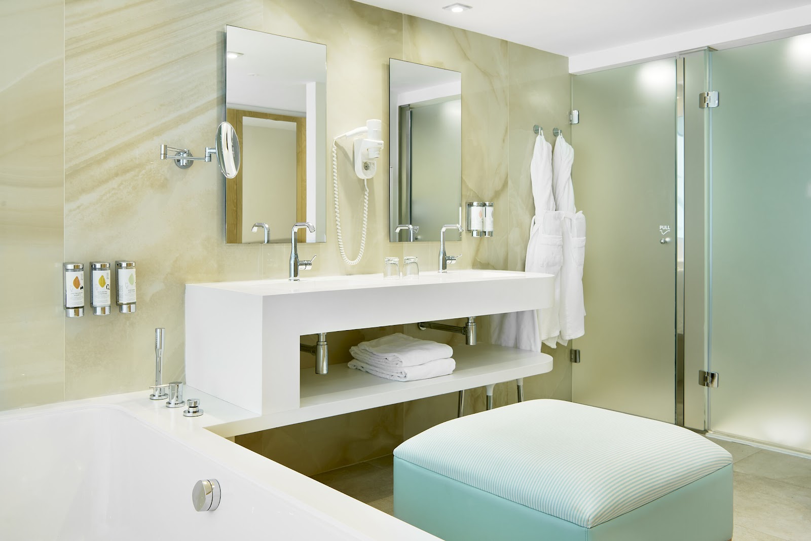 Modern bathroom with white double vanity, cream and green marble walls, glass shower enclosure, white towels, and soaking tub.