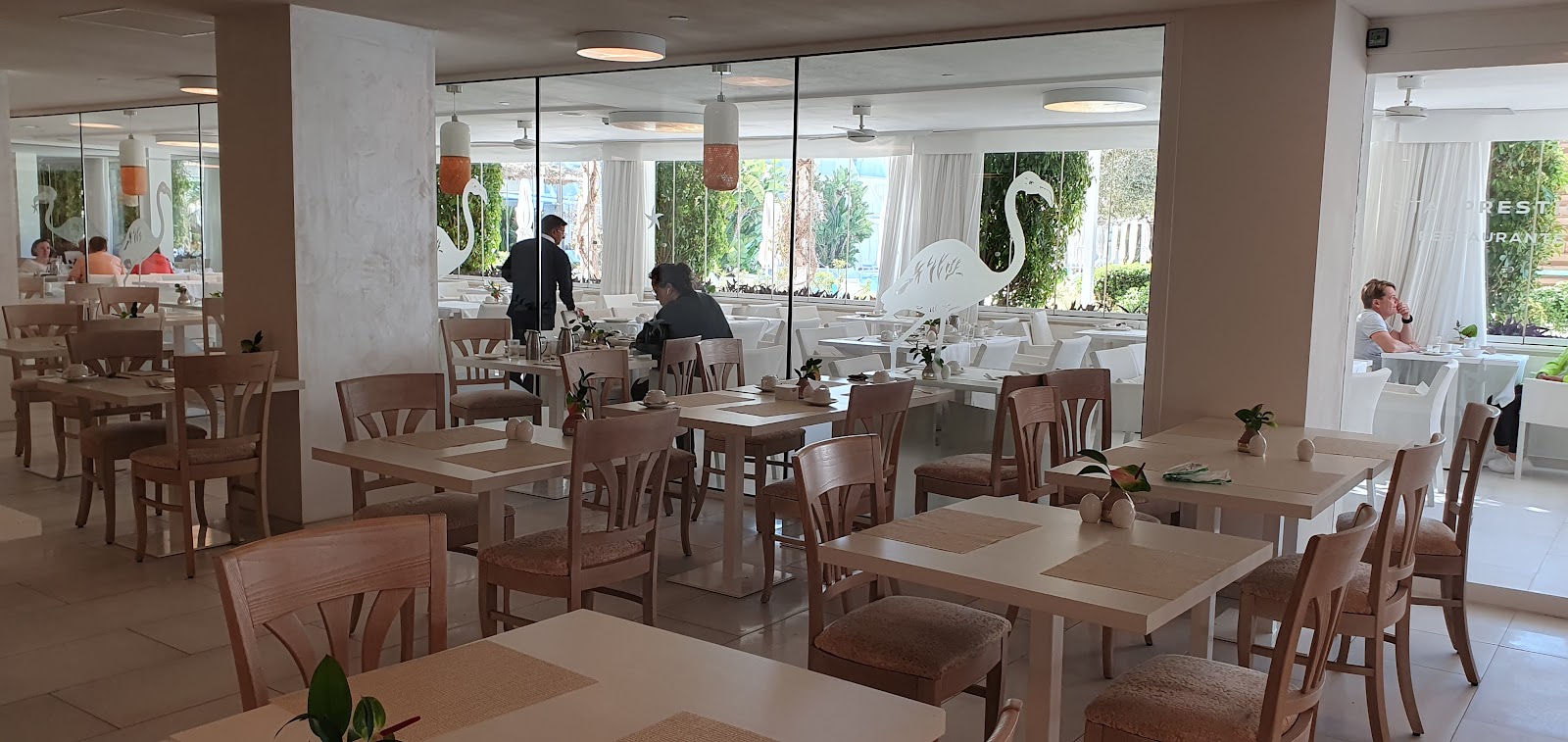 Bright dining room with wooden tables and chairs, large windows overlooking a tropical garden, and a white swan decoration visible through the glass.