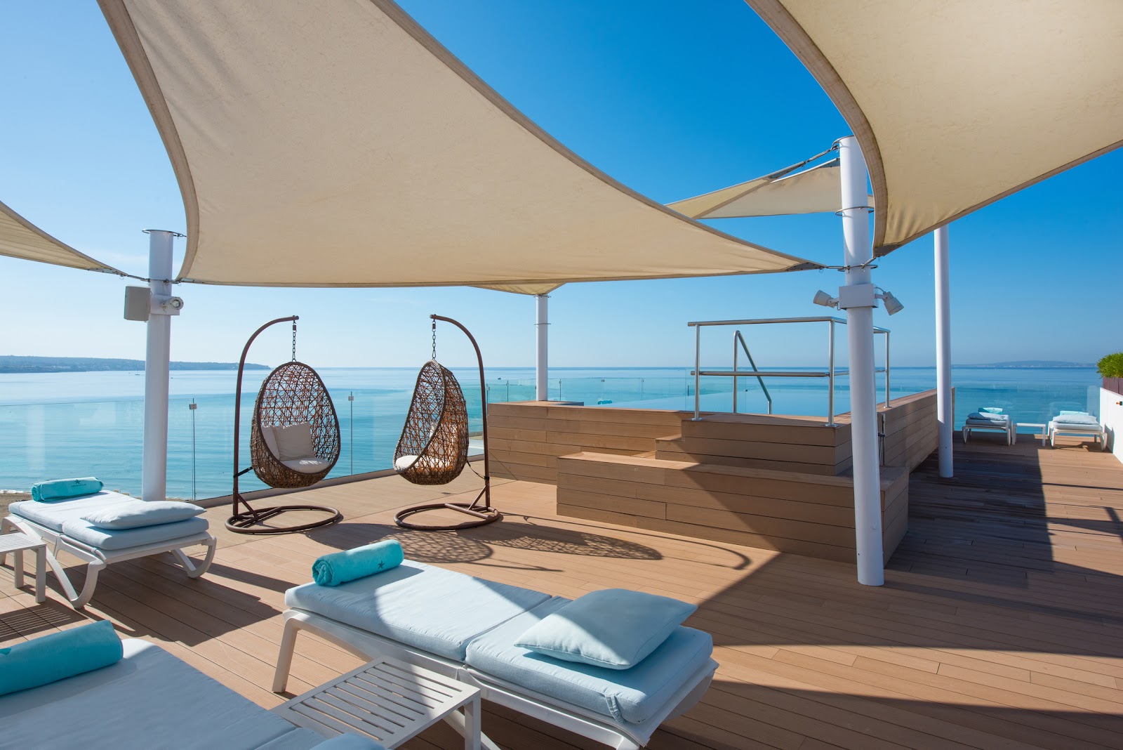 Sunlit pool deck with white umbrellas, turquoise loungers, and a large blue swimming pool, with a modern white resort building visible beyond.