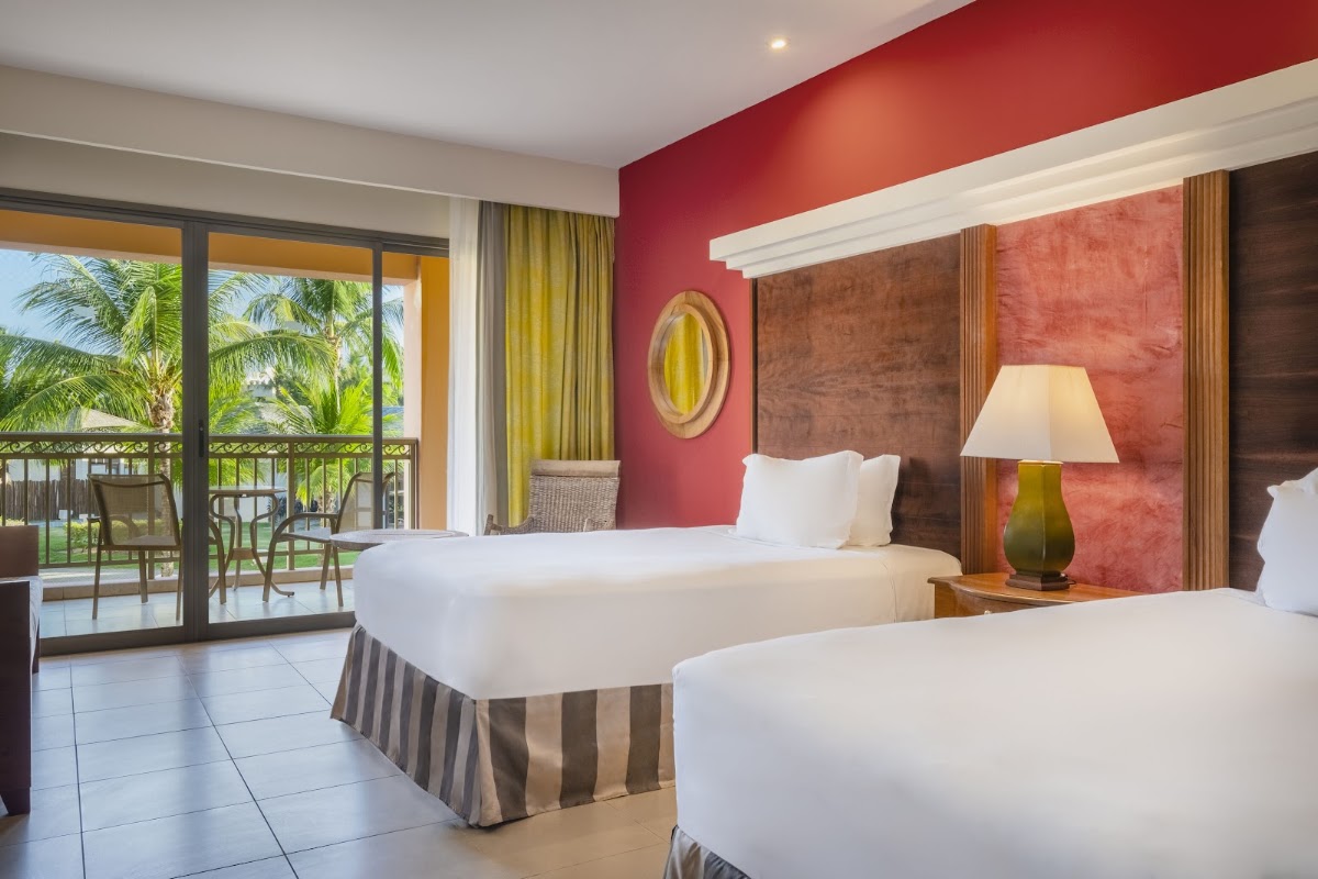 Guest room with two beds, red accent wall, and glass doors opening to a balcony with palm trees and ocean view.