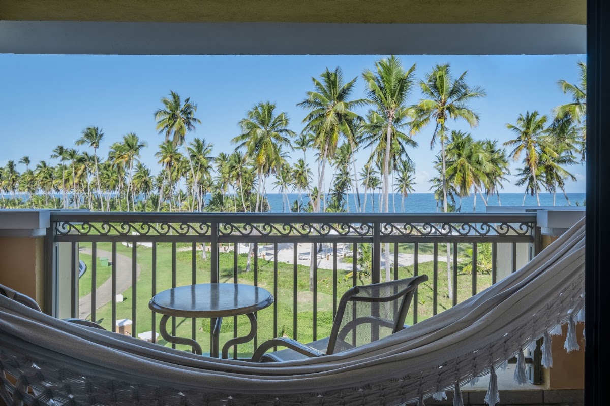 Balcony view from a resort room with a small table and chair, overlooking palm trees, manicured lawn, and ocean beyond.