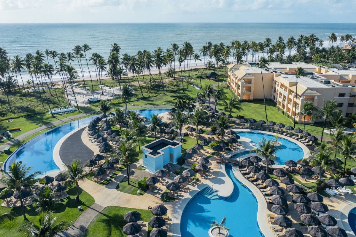 Aerial view of Iberostar Selection Praia do Forte with curved pools, thatched umbrellas, palm trees, beachfront, and tan resort buildings along the ocean.