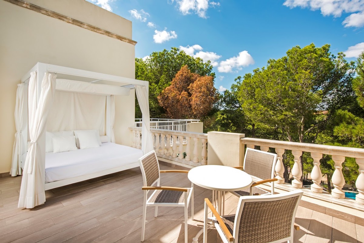 White canopy bed and seating area on a terrace at Iberostar Waves Club Cala Barca, with tree-filled landscape beyond the railing.