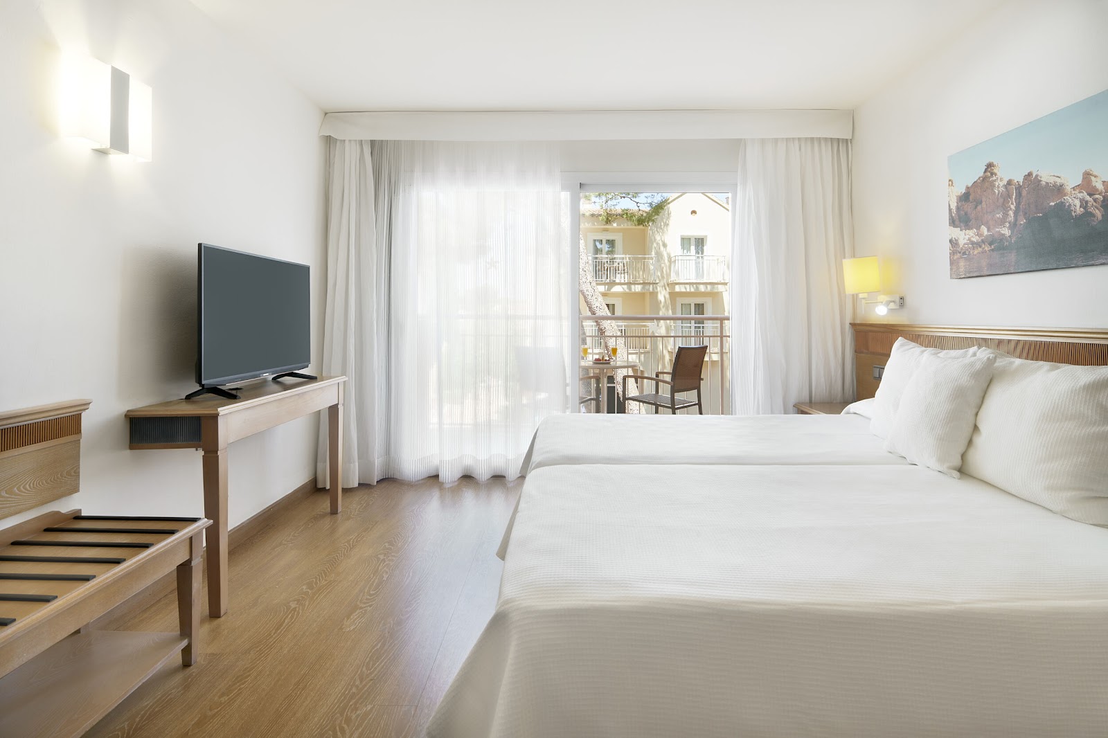 Resort hotel room with double bed, TV, and balcony overlooking palm trees and neighboring buildings.