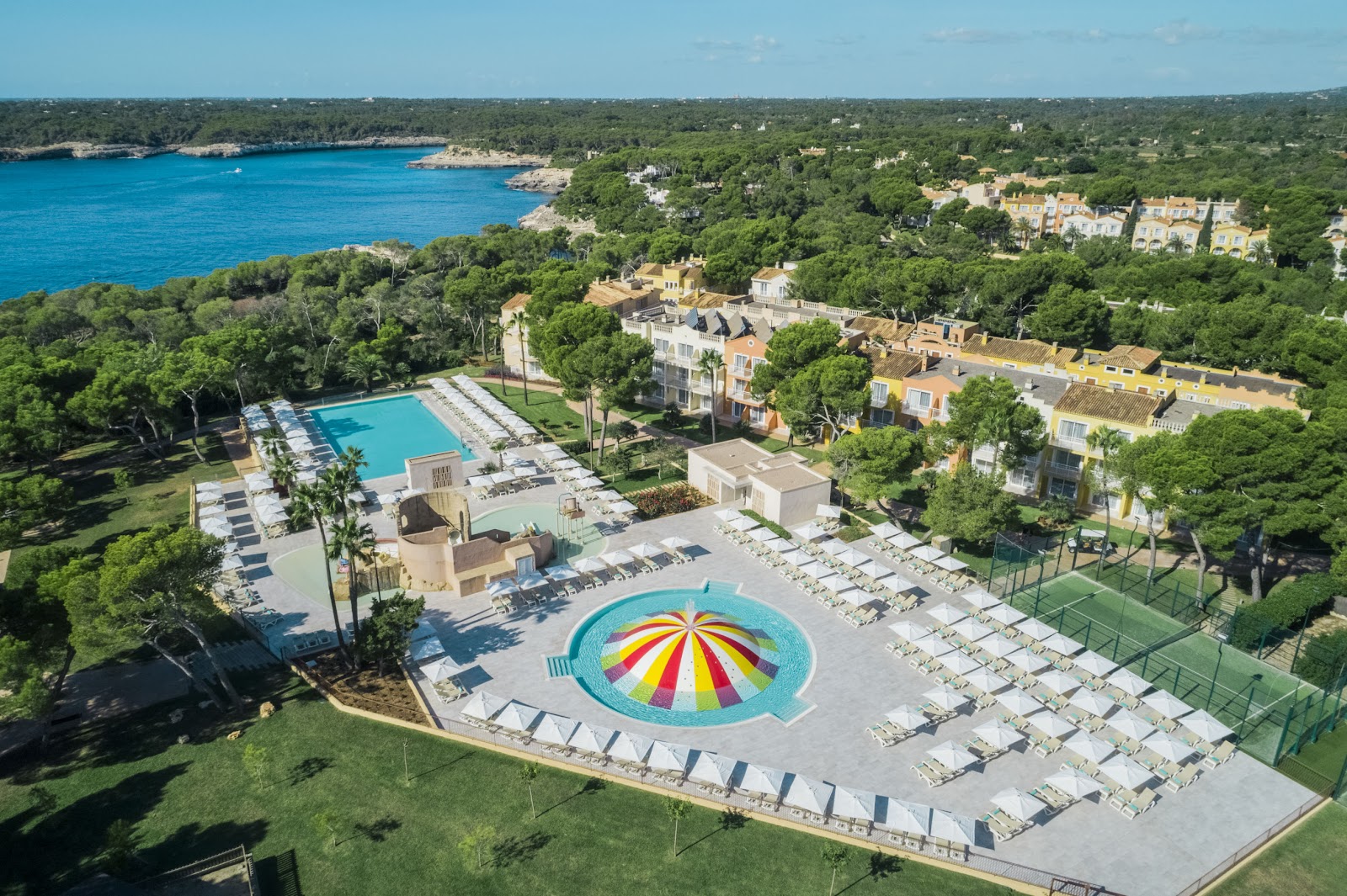 Aerial view of a resort pool complex with a colorful striped umbrella canopy, castle-themed water slide structure, and loungers surrounding shallow turquoise water.