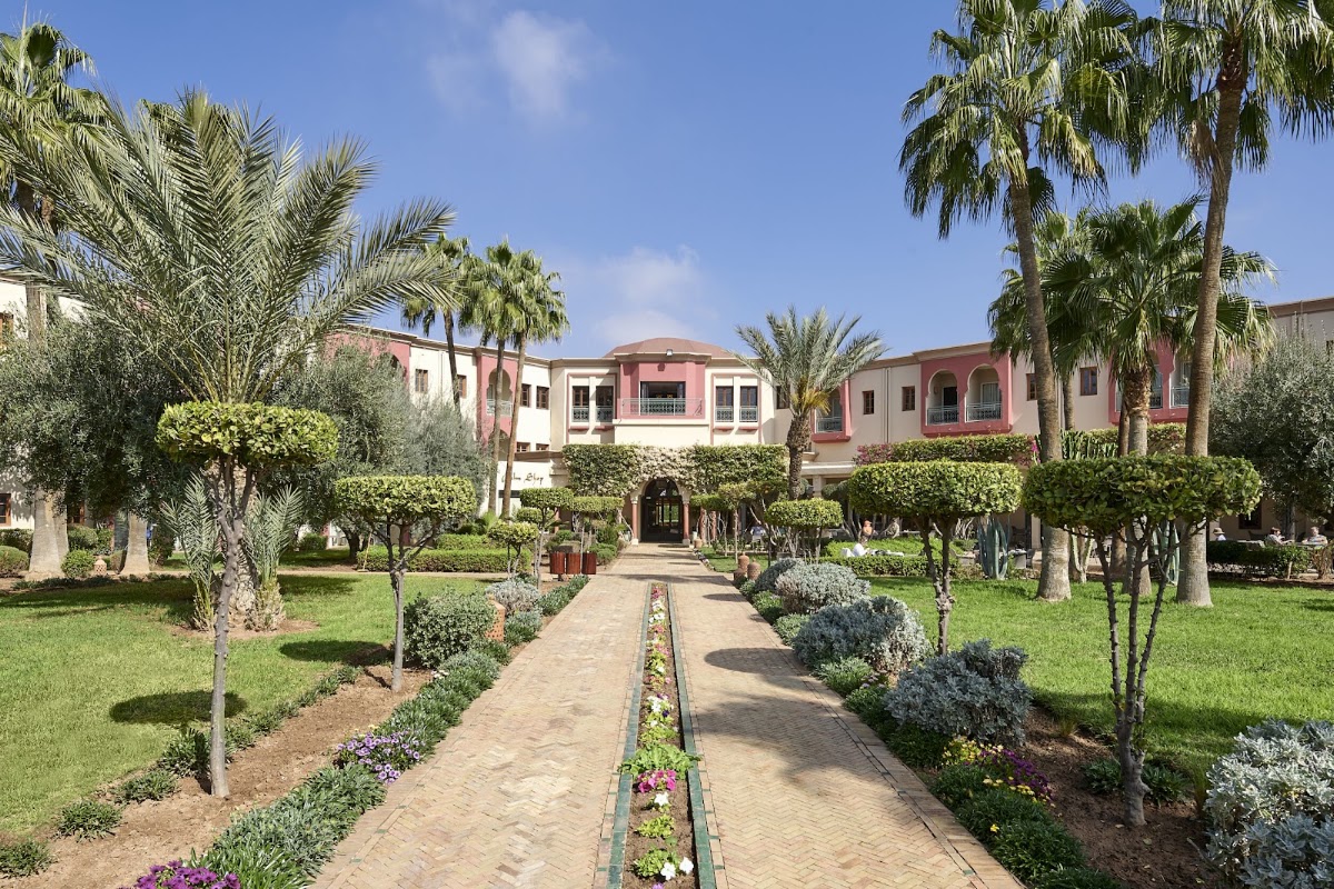 Paved pathway lined with flowering plants and palm trees leading toward pink and white resort buildings under clear blue sky.