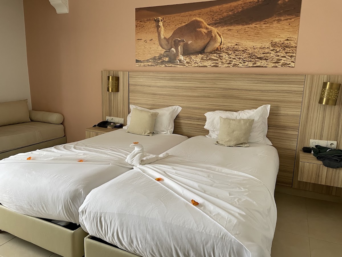 Resort bedroom with two twin beds draped in white linens and decorated with towel animals, wood-paneled headboard, and camel artwork above.