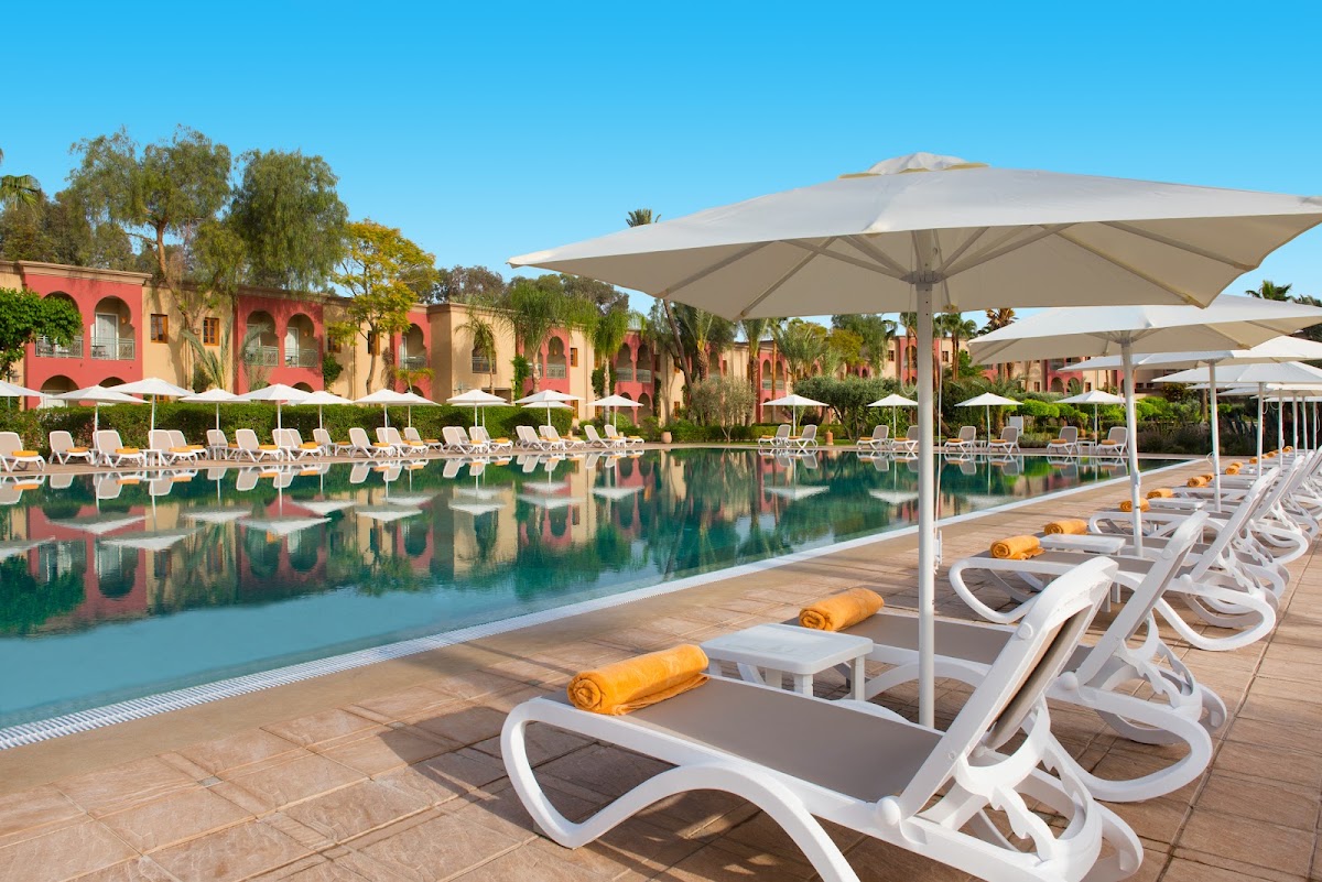 White loungers with yellow towels line a pool deck beside a turquoise pool, with beige umbrellas and red-and-tan buildings beyond under clear blue sky.