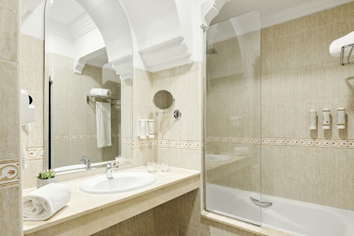 Beige tiled bathroom with large mirror, white oval sink, glass shower enclosure, and rolled towel on counter.
