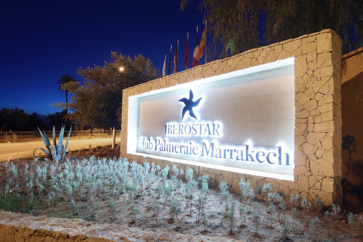 Illuminated Iberostar Club Palmeraie Marrakech sign on a stone wall at dusk, with desert vegetation and flags visible behind it.