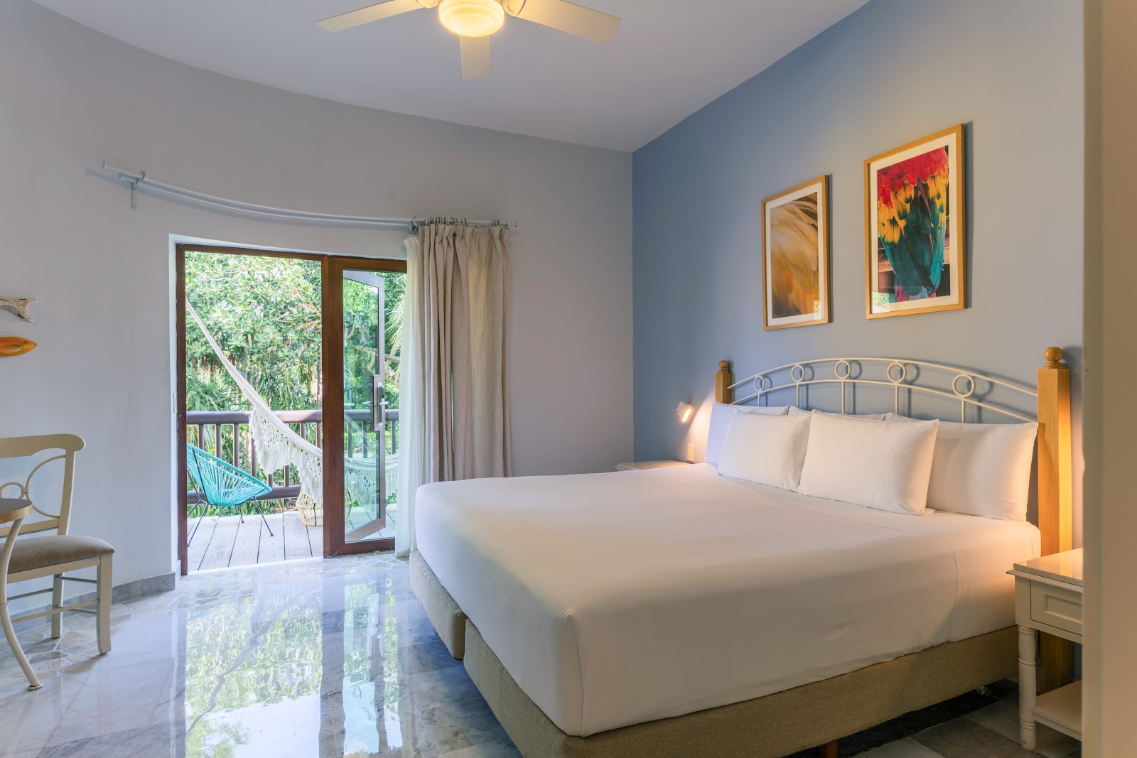 Spacious resort bedroom with wooden four-poster bed, white linens, wood furniture, and sliding glass doors opening to a balcony overlooking tropical grounds.