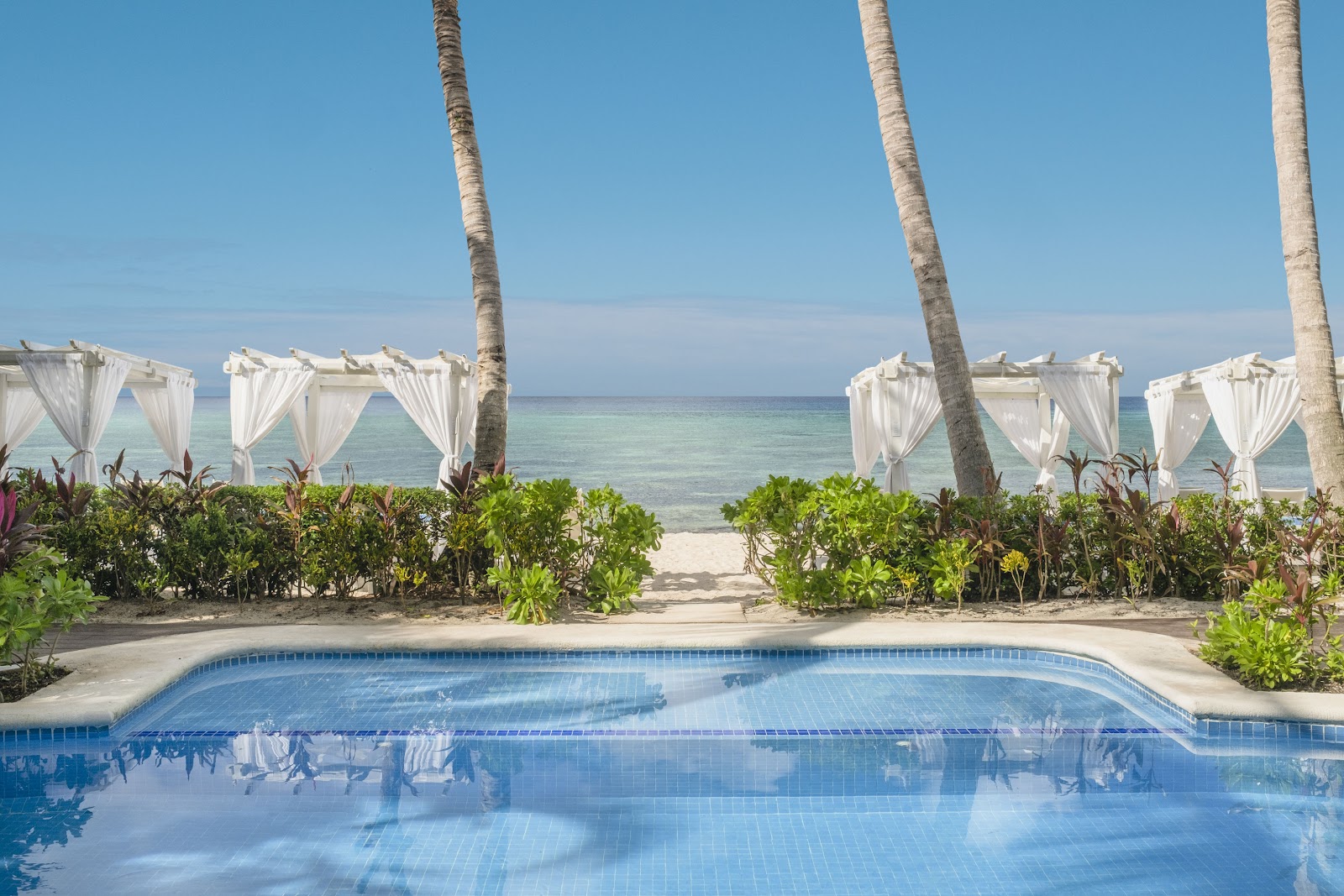 Pool with white cabanas and palm trees overlooking a turquoise ocean beach at Iberostar Waves Cozumel.