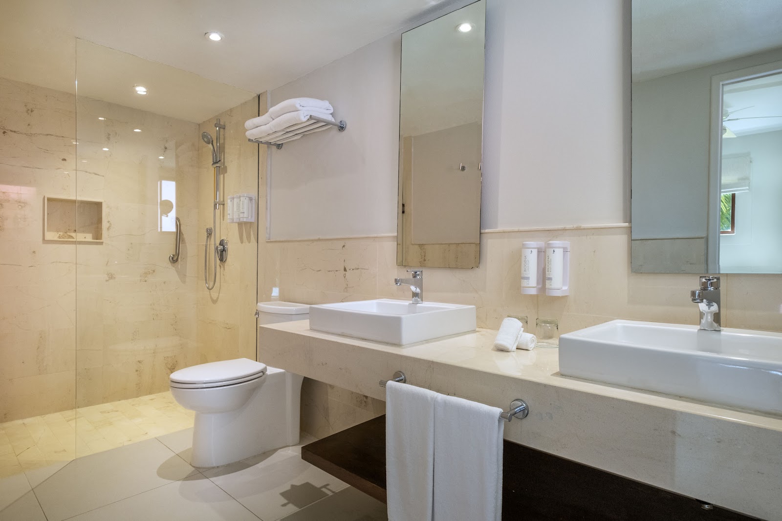 Modern resort bathroom with dual vanities, vessel sinks, walk-in shower, and beige stone tile throughout at Iberostar Waves Cozumel.