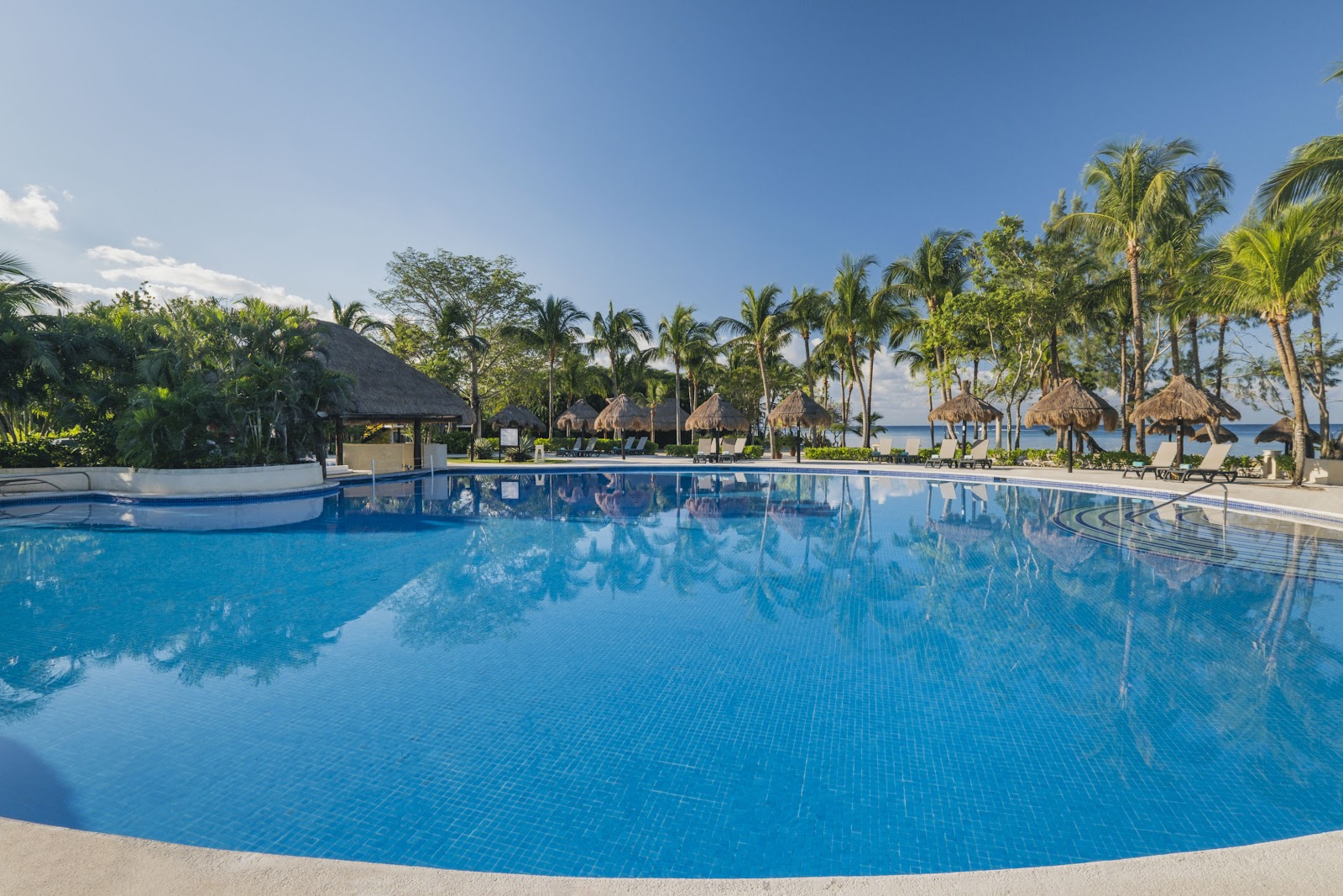 Shaded poolside seating at Iberostar Waves Cozumel with ocean view beyond palm trees and beach cabanas.
