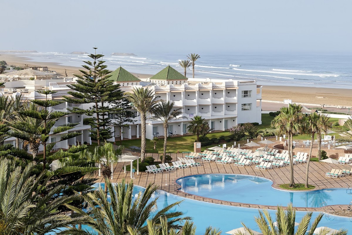 White resort buildings with green-roofed towers overlook a curved pool, palm trees, and sandy beach with ocean waves beyond.