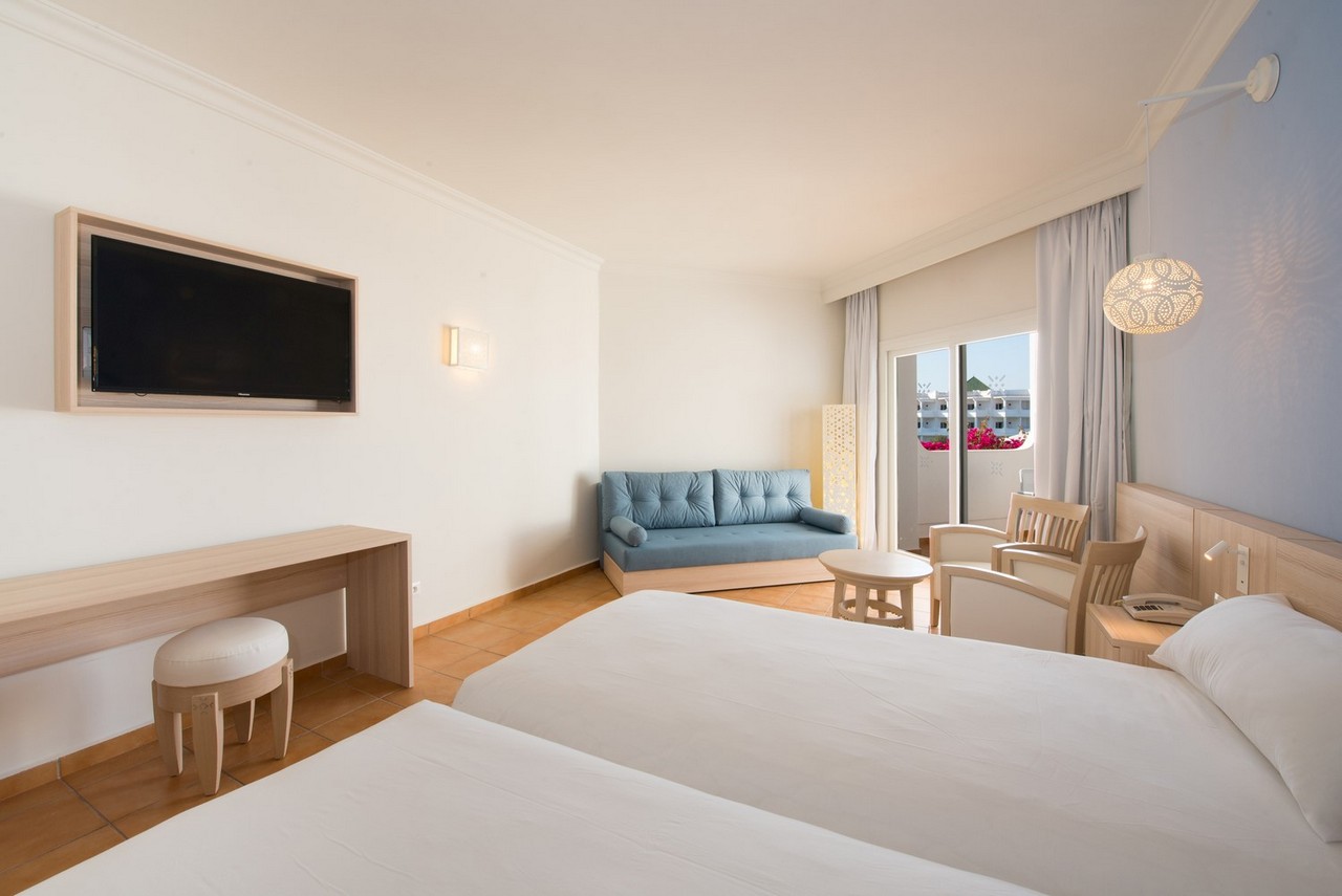 Spacious resort room with two beds, blue sofa, wall-mounted TV, and balcony overlooking grounds with pink flowers.