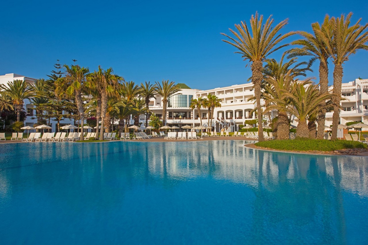 Large turquoise pool lined with palm trees and lounge chairs, white resort building with balconies in background under clear blue sky.