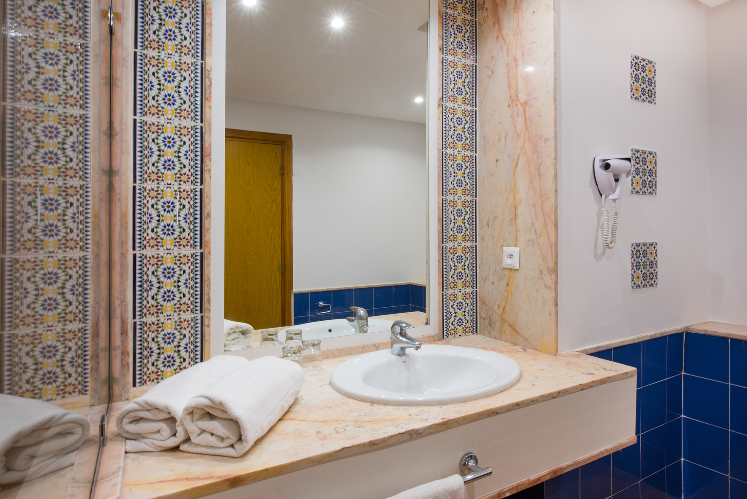 Bathroom with dual white sinks, decorative-framed mirror, marble walls, and glass-enclosed shower at Iberostar Waves Founty Beach.