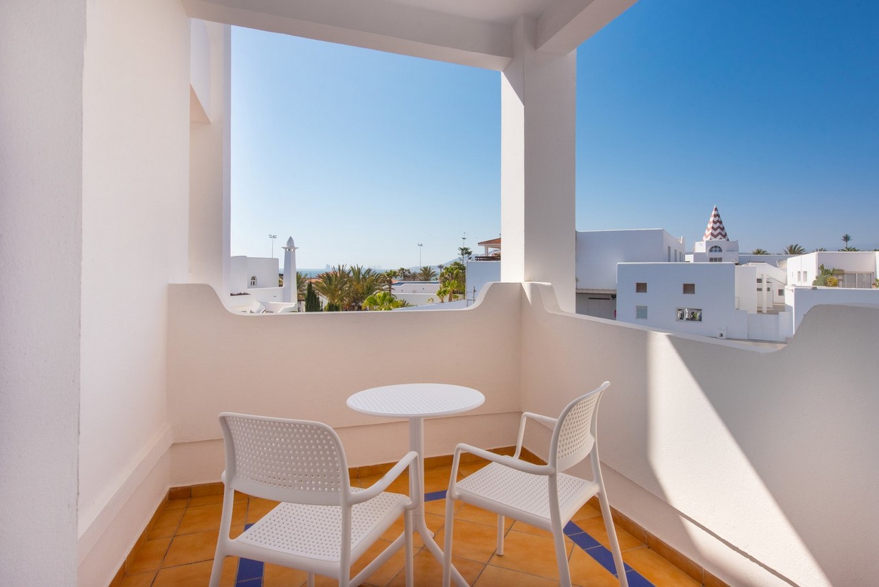 Private balcony with two white chairs and small table overlooking whitewashed resort buildings, palm trees, and minaret in Morocco.