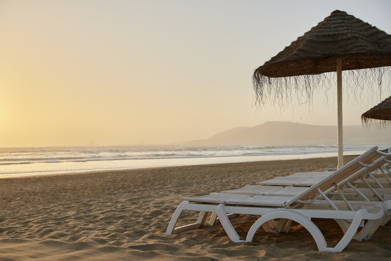 White lounge chairs and thatched umbrellas line the sand at Iberostar Waves Founty Beach at sunrise, with mountains visible across the ocean.