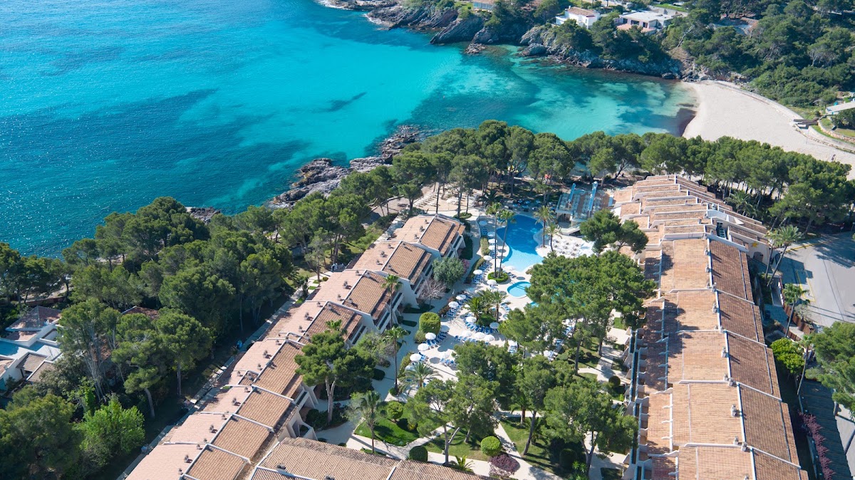 Aerial view of Iberostar Waves Pinos Park with terracotta buildings, pool complex, and turquoise cove nestled between pine-forested cliffs.