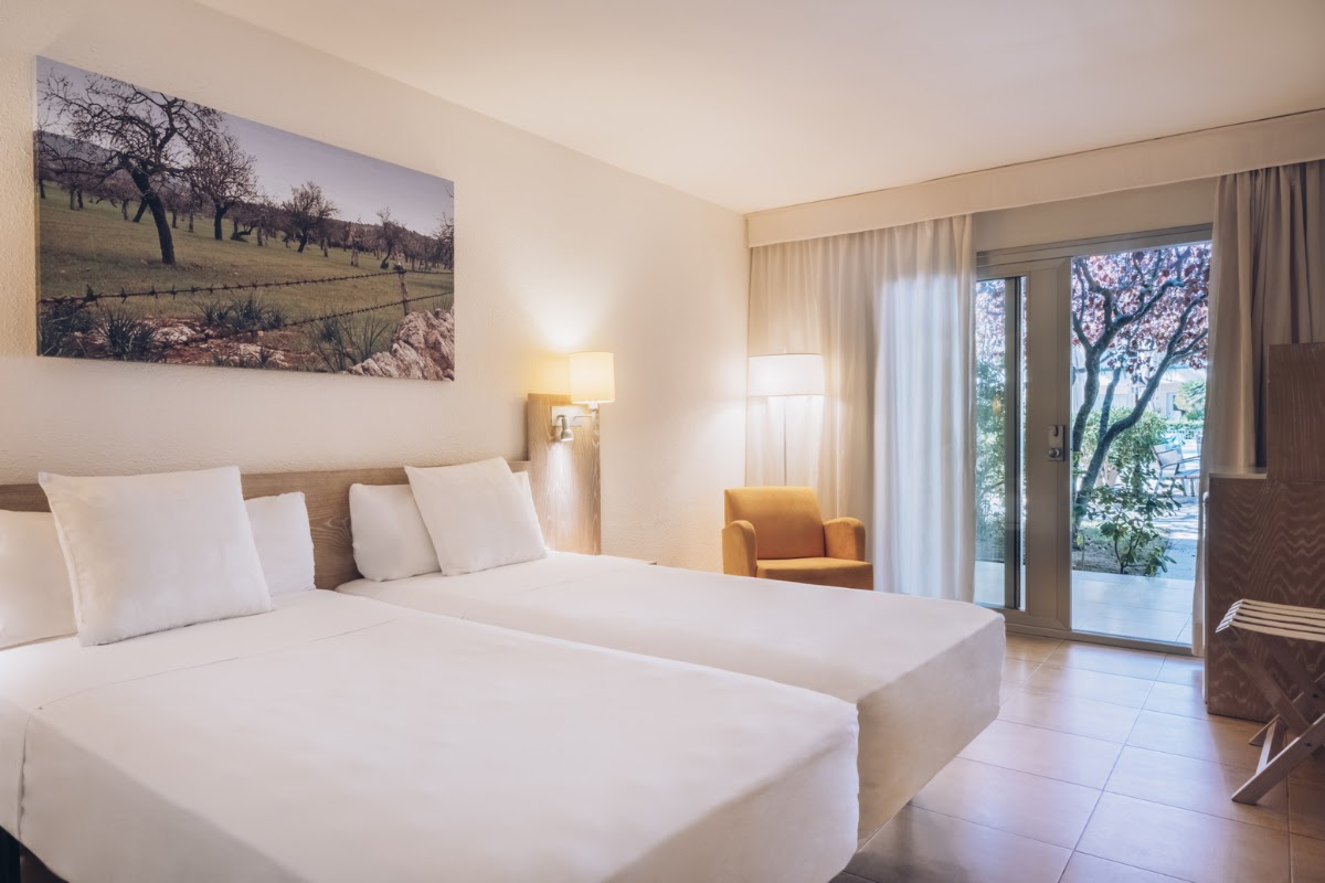 Resort guestroom with two beds, wall art of a rural landscape, and sliding glass doors opening to a terrace with trees.