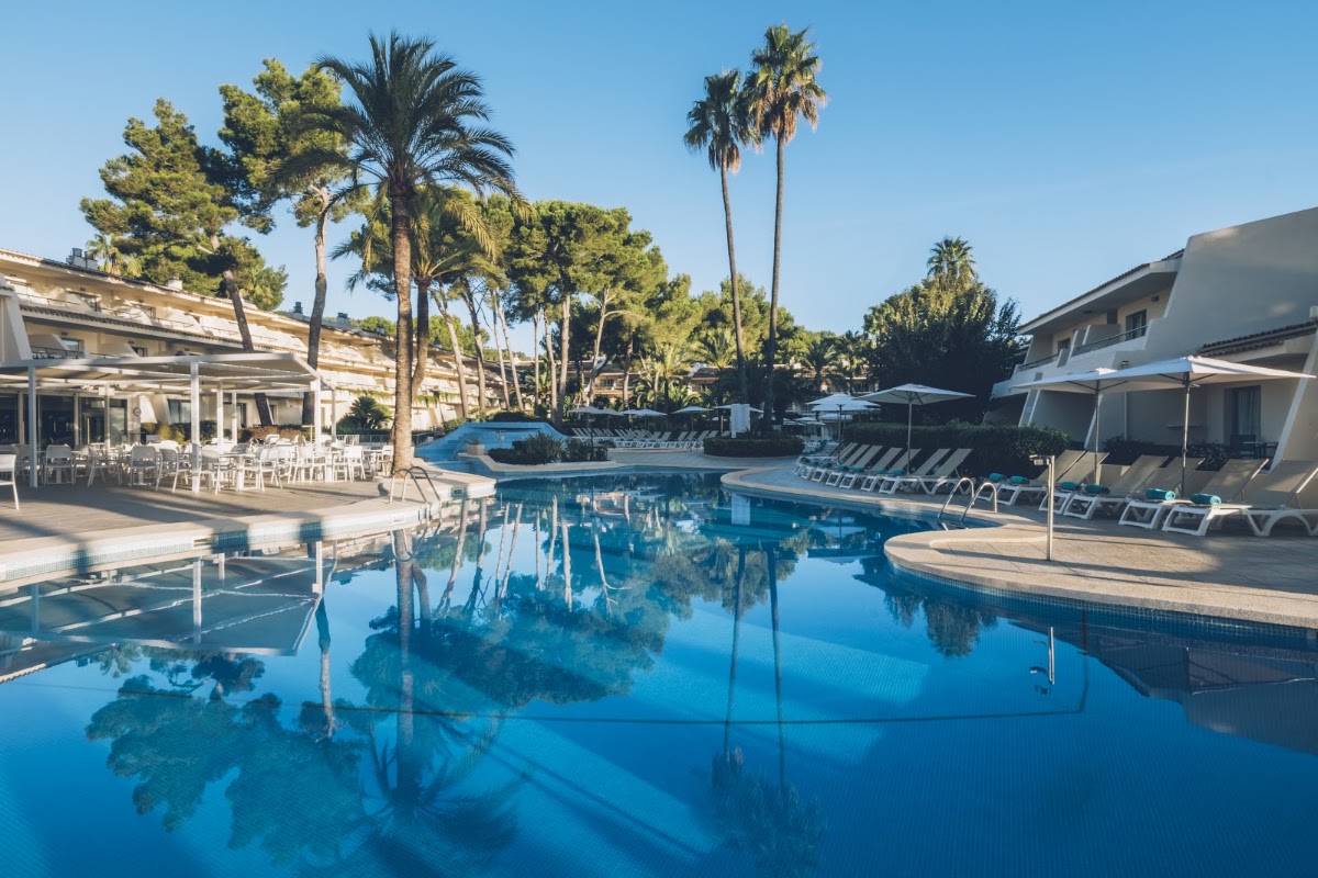 Resort pool with clear blue water, lounge chairs, palm trees, and beige buildings under sunny skies at Iberostar Waves Pinos Park.