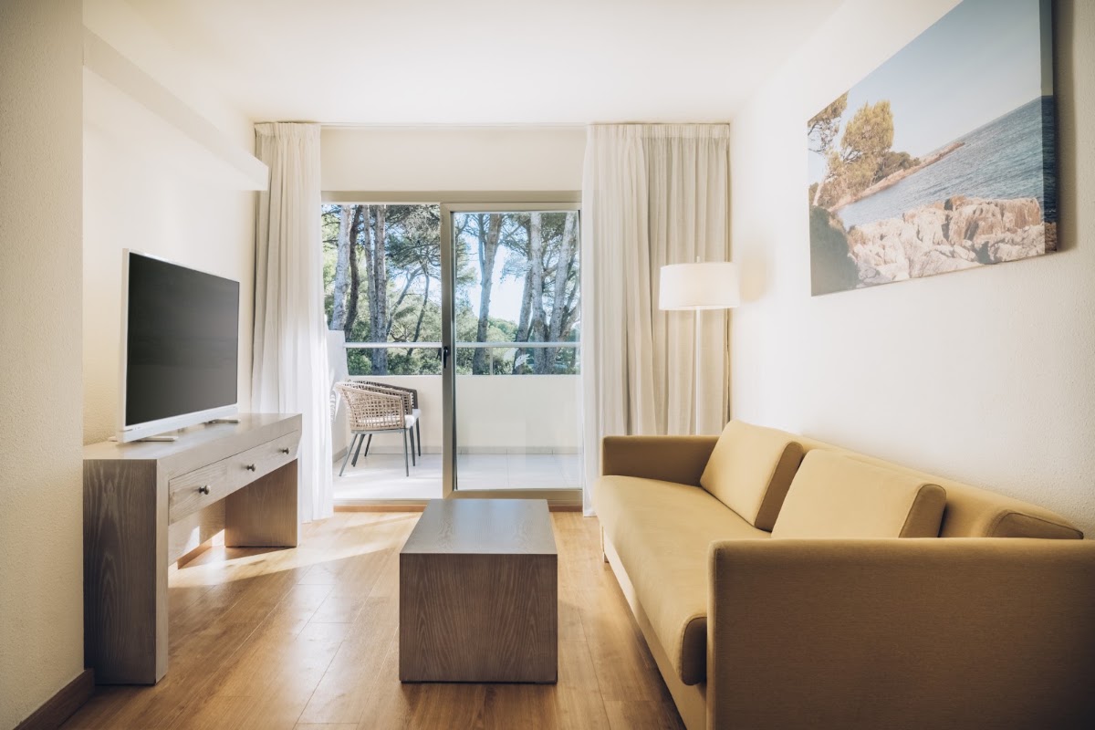 Resort suite with yellow sofa, TV, and glass doors opening to a balcony overlooking pine trees and sea.