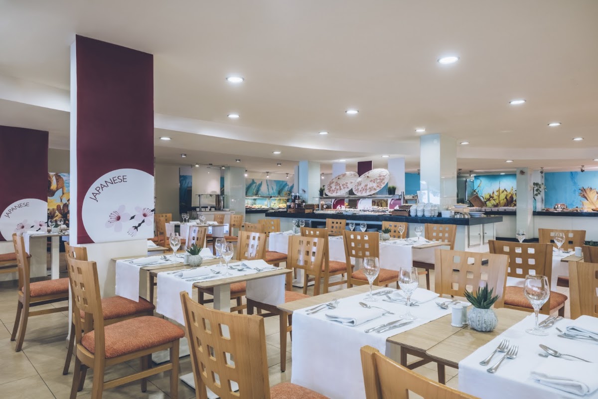 Spacious dining room with wooden chairs, white tablecloths, and an open kitchen at Iberostar Waves Pinos Park, decorated with Japanese-themed signage and artwork.