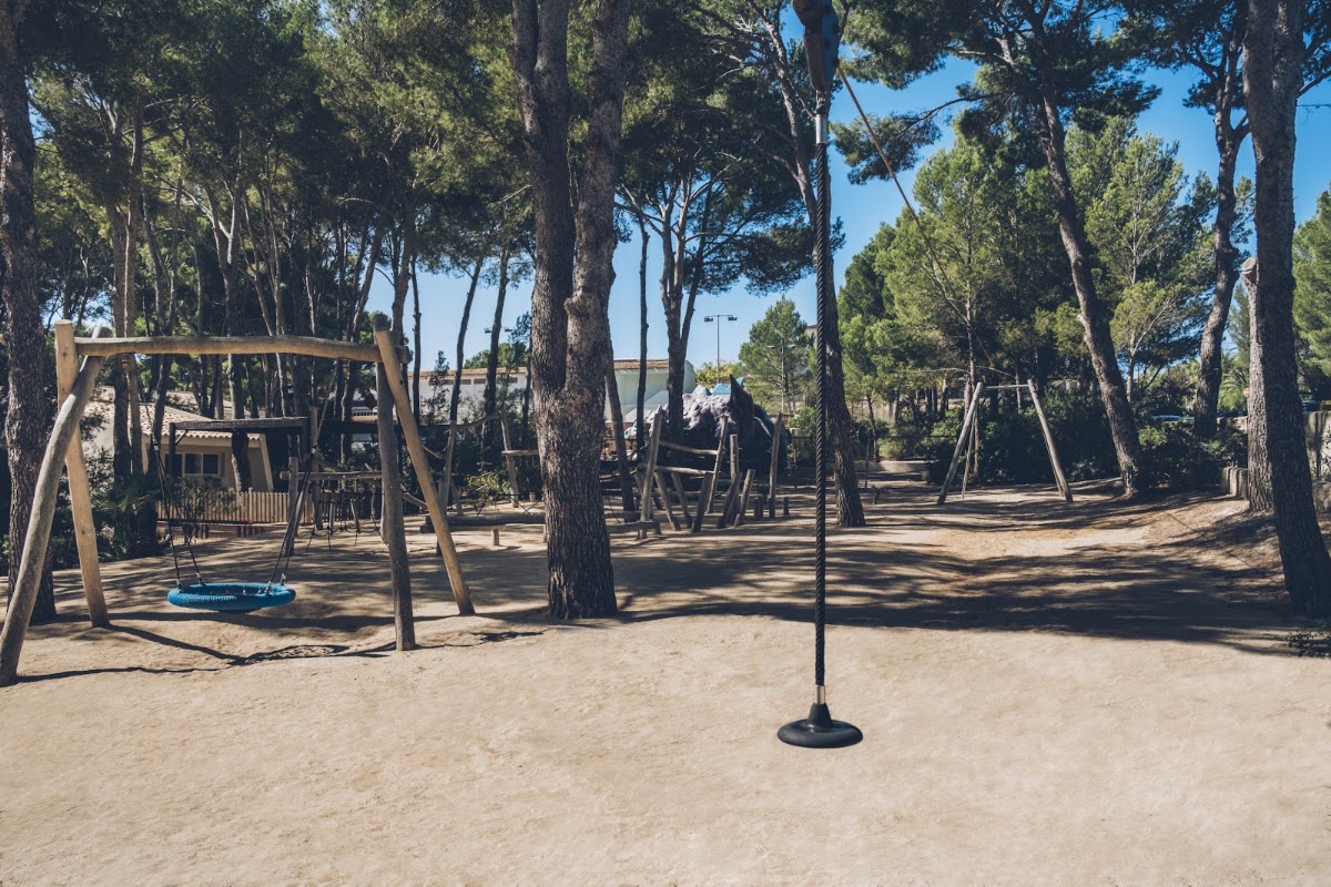 Children's playground with swings and climbing equipment nestled among tall pine trees at Iberostar Waves Pinos Park.