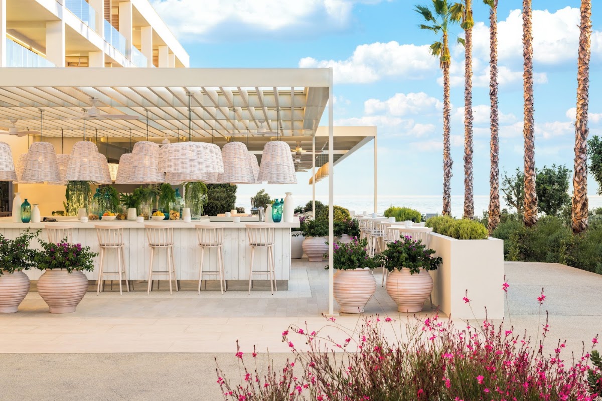 Ikos Andalusia's outdoor bar terrace features woven pendant lamps, bar seating, and tall palm trees with pink flowering plants in foreground.